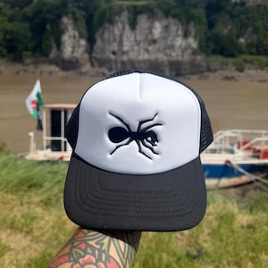May include: A black and white trucker cap featuring a solid white foam front panel with a black embroidered ant graphic, a black brim with horizontal stitching, and a black mesh back panel with an adjustable snapback closure for a secure fit.