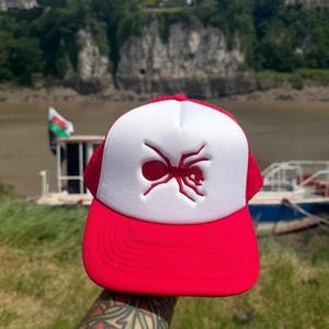 May include: A red and white trucker cap featuring a prominent red embroidered ant graphic centred on the white foam front panel. The cap includes a solid red curved brim and a matching red mesh back panel with an adjustable snapback closure.