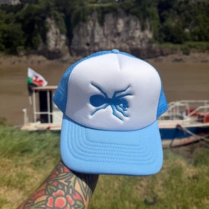 May include: A light blue and white trucker cap featuring a prominent embroidered blue ant graphic on the front foam panel. The cap includes a structured front, a curved brim, and a breathable mesh back section, showcasing a classic design.