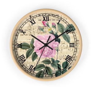 Puede incluir: Un reloj de pared redondo con fondo blanco que presenta una rosa rosa y hojas verdes. La esfera del reloj tiene números romanos y manecillas negras.