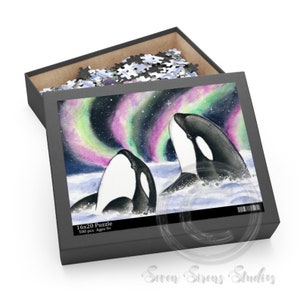 May include: A 500 piece jigsaw puzzle featuring two orcas swimming in the ocean under a vibrant aurora borealis. The puzzle box is black with white text that reads "16x20 Puzzle 500 pcs Ages 9+"