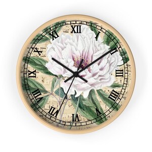 Puede incluir: Una flor de peonía blanca y rosa con hojas verdes sobre un fondo de partitura musical vintage. La flor está centrada en una esfera de reloj redonda de madera con marcas de números romanos.