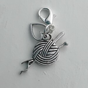 May include: A silver charm depicting a ball of yarn with knitting needles and a heart-shaped detail. It has a lobster clasp for attaching to a bracelet or necklace. Perfect for craft enthusiasts and knitters.