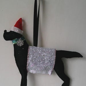 May include: A black dog ornament featuring a red and white Santa hat, a green glittery collar with a silver snowflake charm, and a white glittery blanket draped over its back. The ornament hangs from a black ribbon loop for display purposes.