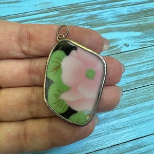 May include: A pendant with a floral design. The pendant features a pink flower with green leaves and black accents, set within a silver-colored frame. The pendant is held by a silver-colored loop.