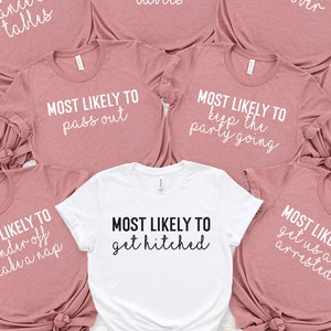 May include: Nine pink t-shirts with white text that says "Most Likely To" followed by a different phrase on each shirt. The phrases are: "Dance on Tables", "Bring Home a Stray Dog", "Dance on Tables", "Be Sober", "Pop Bottles", "Pass Out", "Keep the Party Going", "Wander Off to Take a Nap", "Get Us All Arrested". One white t-shirt with black text that says "Most Likely To Get Hitched".