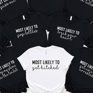 May include: Nine black t-shirts with white text that says "Most Likely To" followed by a different phrase on each shirt. The phrases are: "Kiss a Stranger", "Pass Out", "Lose My Shorts", "Dance on Tables", "Snapchat the Whole Night", "Pop Bottles", "Break Your Heart", "Drop My Phone in the Toilet", "Get Us All Arrested". One white t-shirt with black text that says "Most Likely To Get Hitched".