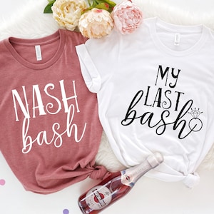 May include: Two t-shirts, one dusty rose and one white, are displayed. The dusty rose shirt reads "NASH bash" in white script. The white shirt says "MY LAST bash" with a diamond ring graphic. A bottle of sparkling wine is visible.