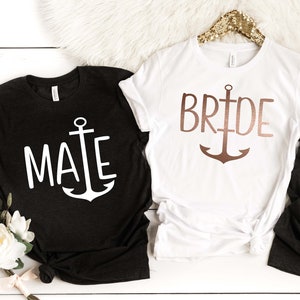 May include: Two t-shirts with an anchor design. One black t-shirt says "Mate" in white text with a white anchor. The other white t-shirt says "Bride" in rose gold text with a rose gold anchor.