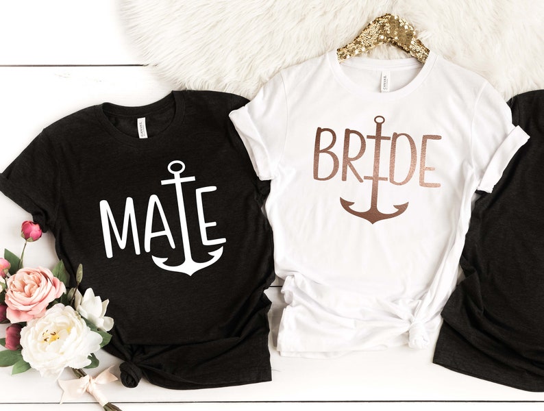 May include: Two t-shirts with an anchor design. One black t-shirt says "Mate" in white text with a white anchor. The other white t-shirt says "Bride" in rose gold text with a rose gold anchor.