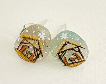 May include: A pair of silver stud earrings with a wintery scene of a stable with a star painted on each earring. The stable is painted in shades of brown and the background is a light blue with white snowflakes.