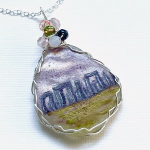 May include: A silver chain necklace with a teardrop-shaped pendant featuring a painted image of Stonehenge. The pendant is surrounded by a silver wire frame and accented with colorful beads.