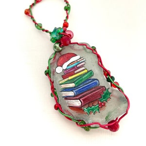 May include: A Christmas ornament made from a clear glass rock with a painted image of a stack of books wearing a Santa hat and holly. The rock is wrapped in red and green wire with beads.