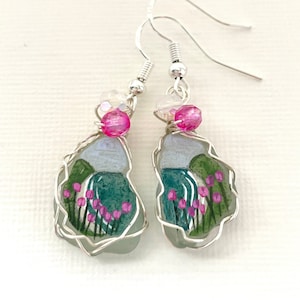 May include: Silver dangle earrings with a painted seascape scene inside a clear glass teardrop shape. The scene features pink flowers, green grass, and a blue ocean. The earrings are accented with pink and clear crystal beads.