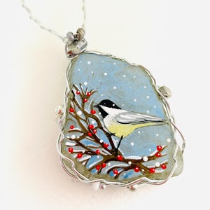 Puede incluir: Un adorno de vidrio pintado a mano que representa un carbonero posado en una rama con bayas rojas. El adorno está rodeado por un marco de alambre plateado y tiene un lazo para colgar.