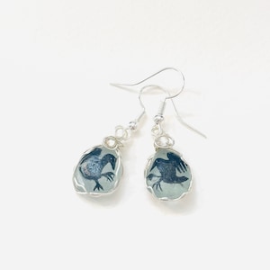 May include: Silver dangle earrings with a clear glass teardrop shaped charm featuring a black silhouette of a bird.