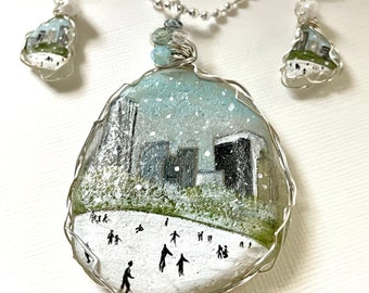 May include: A silver necklace and earrings set with a winter scene painted on a white stone. The scene depicts a snowy city with people ice skating on a frozen lake. The necklace and earrings are both made with silver wire and have a delicate, whimsical feel.