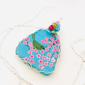 Può includere: Una collana con catena d'argento con un ciondolo in vetro blu che presenta un uccello verde dipinto a mano appollaiato su un ramo di fiori rosa. Il ciondolo è avvolto in filo d'argento e sormontato da perline colorate.