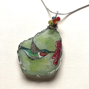 May include: A silver chain necklace with a green sea glass pendant featuring a hand-painted hummingbird in flight, drinking nectar from a red flower. The pendant is wrapped in silver wire and accented with colorful beads.