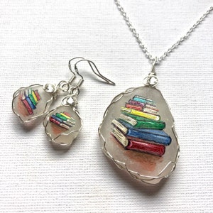 May include: A silver necklace and earrings set featuring a hand-painted glass pendant with a stack of colorful books. The pendant is surrounded by silver wire.