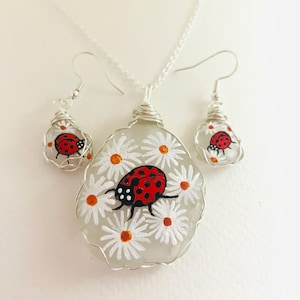 以下が含まれることがあります： 手作りのジュエリーセット。ペンダントネックレスとイヤリングのセットです。白い背景にデイジーの花とてんとう虫のデザインが施されています。てんとう虫は赤色で黒い斑点があり、イヤリングとペンダントはワイヤーで包まれています。