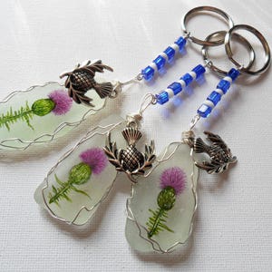 May include: Three keychains featuring a pressed thistle flower encased in clear glass with a silver thistle charm. Each keychain has a blue and white beaded chain.