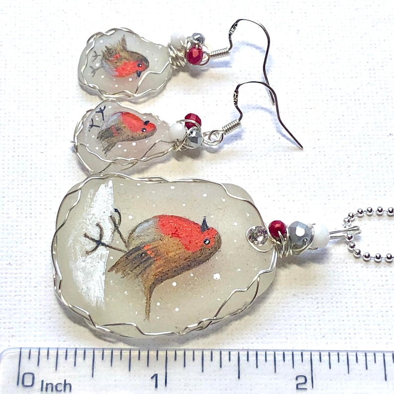 May include: A set of three robin bird ornaments, each with a different pose, painted on a white glass background. The ornaments are wrapped in silver wire and have red and silver beads. The largest ornament is about 2 inches long.