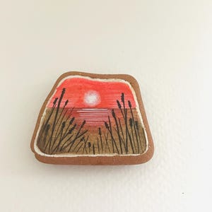 Puede incluir: Piedra pintada a mano con una escena de puesta de sol. La piedra está pintada con una puesta de sol roja y naranja sobre una masa de agua con siluetas negras de hierba alta en primer plano.