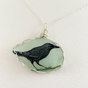 May include: A handcrafted pendant necklace featuring a black bird painted on a piece of sea glass. The sea glass is a light blue-green color, and the bird is depicted with fine detail. The pendant is wrapped in silver wire and hangs from a delicate silver chain.