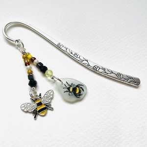 Könnte beinhalten: Silbernes Lesezeichen mit floralem Design und einem Anhänger mit einer gemalten Biene auf einem klaren Glasstück. Der Anhänger ist an einer Perlenkette mit schwarzen, weißen und gelben Perlen befestigt.