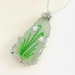 Peut inclure: Un pendentif en fil d'argent enveloppé avec une pièce de verre de mer verte avec des fleurs de perce-neige blanches peintes à la main et des feuilles vertes.