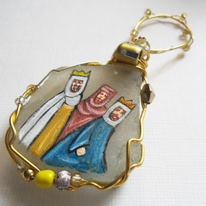 May include: A gold wire-wrapped ornament featuring a painted image of the three wise men on a piece of sea glass. The ornament is decorated with gold wire, beads, and a yellow bead.