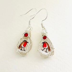 Puede incluir: Un par de pendientes de alambre plateado con un encanto de vidrio blanco que presenta un petirrojo pintado a mano sobre un fondo nevado. Cada encanto tiene una cuenta roja y una cuenta de perla blanca.