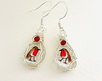 May include: A pair of silver wire wrapped earrings with a white glass charm featuring a hand-painted robin on a snowy background. Each charm has a red bead and a white pearl bead.