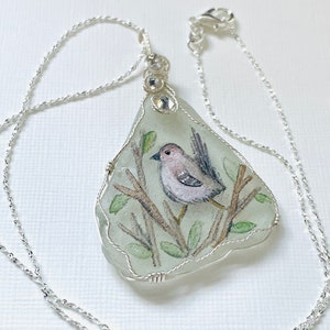 Puede incluir: Un collar de cadena de plata con un colgante que presenta un pájaro pintado a mano en una rama, encerrado en vidrio transparente y adornado con diamantes de imitación.
