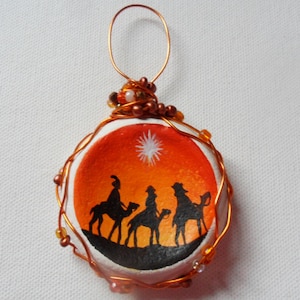 May include: A round, white ornament with a wire frame and orange and brown beads. The ornament is painted with a scene of three wise men riding camels towards a star in the sky.