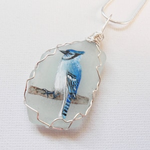 May include: A blue jay painted on a piece of sea glass, framed with silver wire, and hanging from a silver chain.