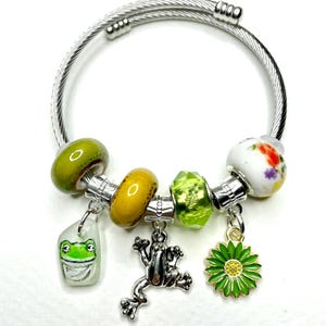 Könnte beinhalten: Ein silberfarbenes Armband mit verschiedenen Anhängern. Die Anhänger umfassen grüne und gelbe Perlen, einen Frosch-Anhänger, einen grünen Blumen-Anhänger und einen Anhänger mit einem Froschgesicht. Das Armband hat ein Kabeldraht-Design.