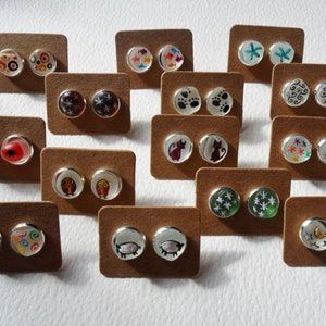 Può includere: Assortimento di orecchini fatti a mano con vari disegni, tra cui gufi, gatti, stelle, fiocchi di neve e impronte di zampe. Gli orecchini sono esposti su piccole carte marroni.