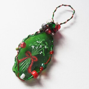 Puede incluir: Un adorno de vidrio verde con un diseño floral pintado y una cinta roja, rodeado de cuentas rojas y verdes y alambre.