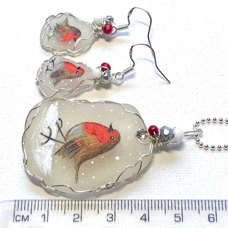 May include: A set of three robin bird jewelry pieces, each featuring a hand-painted robin on a white background with red accents. The necklace is a larger piece with a silver wire wrapped around the edge. The earrings are smaller and have silver hooks. The pieces are all approximately 2 inches in length.