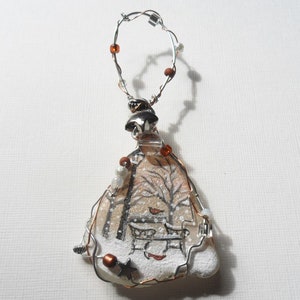 May include: A handcrafted winter scene ornament. The ornament depicts a painted winter landscape with bare trees, a bench, and falling snow. It is framed with silver wire and decorated with beads, and has a silver wire loop for hanging.
