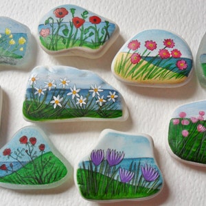 Pode incluir: Uma coleção de peças de vidro do mar pintadas à mão, cada uma retratando uma cena costeira diferente. A obra de arte inclui flores coloridas, grama verde, água azul e um céu azul claro. As peças variam em forma e tamanho.