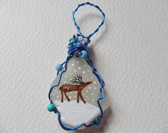 May include: A white sea glass ornament with a hand-painted reindeer in a snowy scene. The ornament is wrapped in blue wire and accented with blue and turquoise beads.