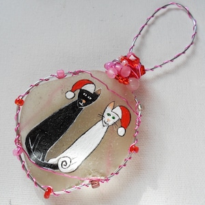 May include: A Christmas bauble featuring two cats wearing Santa hats, painted on a white, round, glass stone. The bauble is decorated with pink and red beads and a wire hanger.