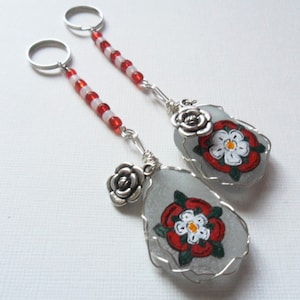 Puede incluir: Dos llaveros plateados con cuentas rojas y blancas. Cada llavero tiene un dije plateado de una rosa y un dije de vidrio blanco con una rosa roja y blanca pintada.