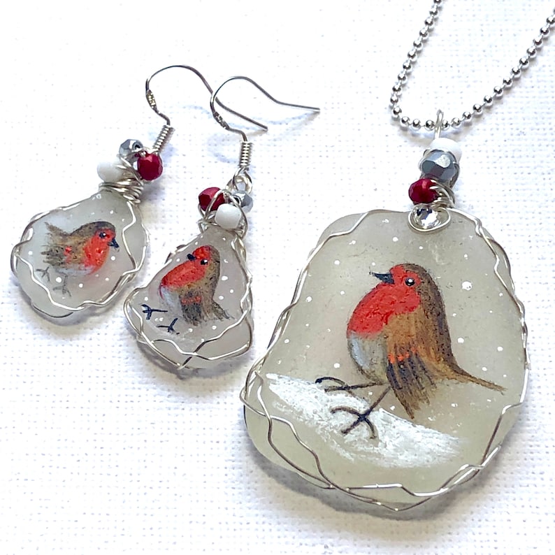 May include: A set of robin bird jewelry featuring a necklace and earrings. The robin is painted on a white sea glass pendant and earrings, with silver wire wrapping. The necklace has a silver chain and the earrings have silver hooks.