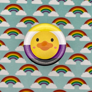 May include: A yellow, purple, and black button with a cartoon duck face. The duck has a yellow beak and orange feet. The button is on a background of rainbow clouds.