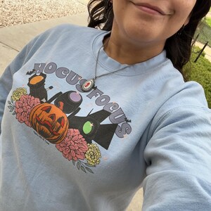 May include: A light blue sweatshirt with a graphic print of the Sanderson sisters from the movie Hocus Pocus. The graphic includes a pumpkin, flowers, and the text "Hocus Focus".