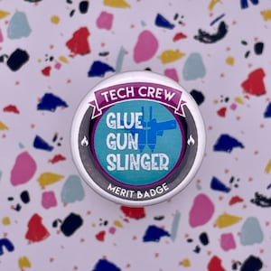 May include: A silver button with a blue and white design. The button says "Tech Crew Glue Gun Slinger Merit Badge".
